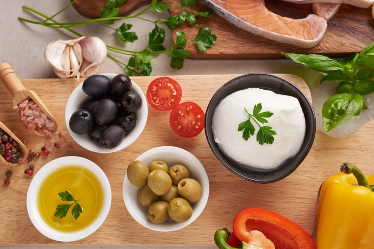 An assortment of Mediterranean ingredients including black and green olives, olive oil, tomatoes, bell peppers, garlic, salmon