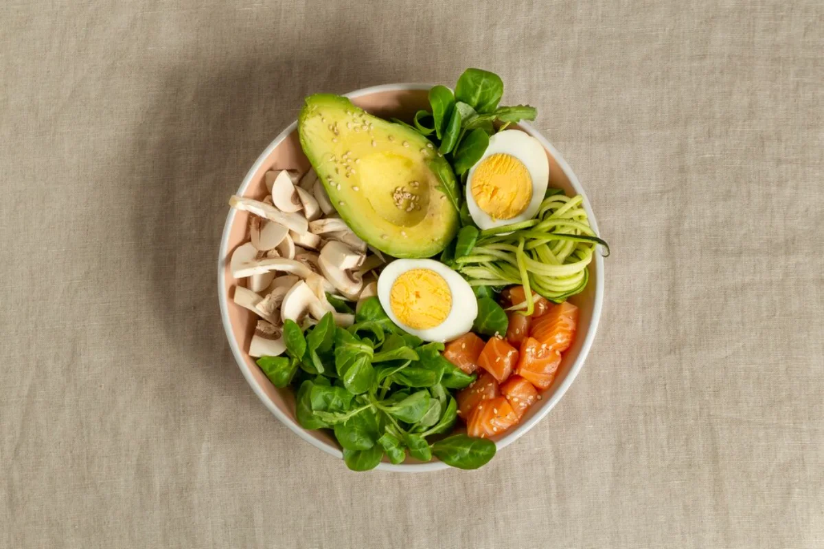 A nutritious bowl of salad containing half an avocado with sesame seeds, boiled eggs, sliced mushrooms, spinach leaves