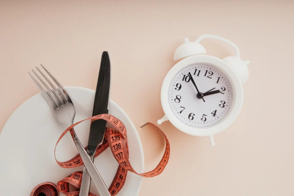 A white plate with a fork, knife, and measuring tape coiled on it, next to a white alarm clock