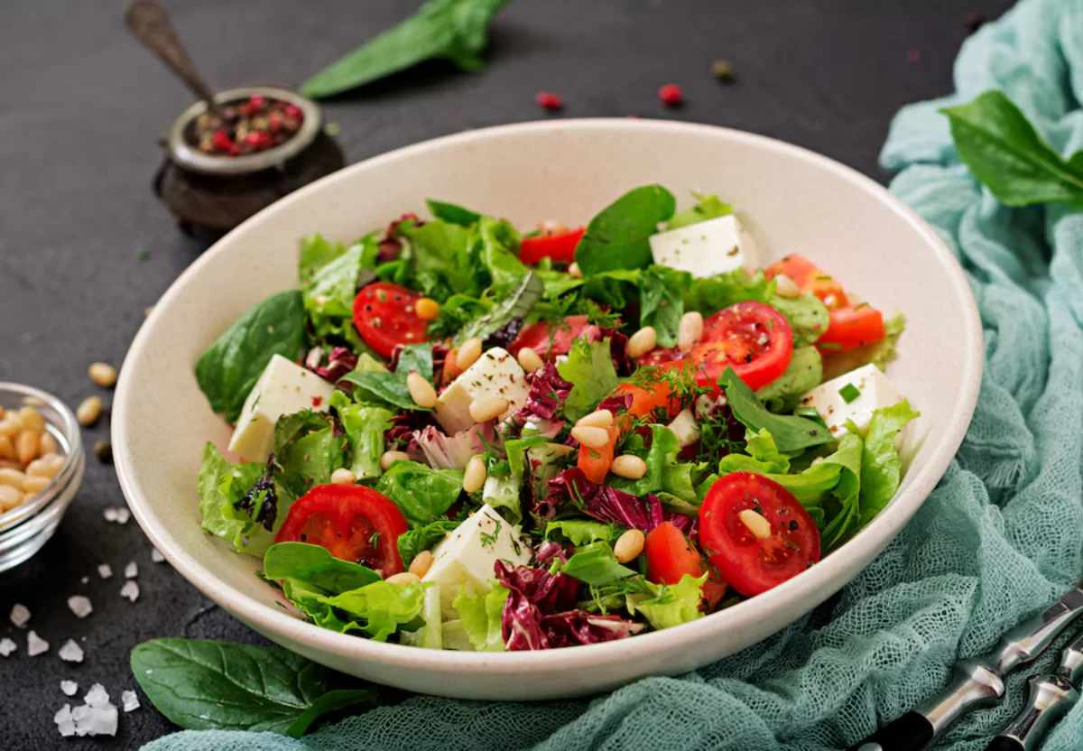 A fresh mixed salad in a bowl featuring leafy greens, sliced tomatoes, cubes of cheese, and pine nuts