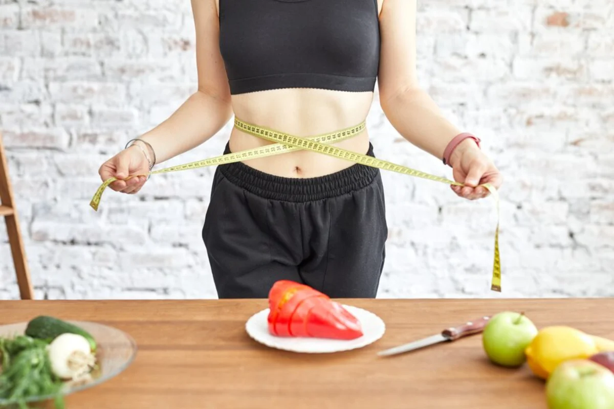 A woman measuring her waist with a tape measure in front of a table with healthy foods 