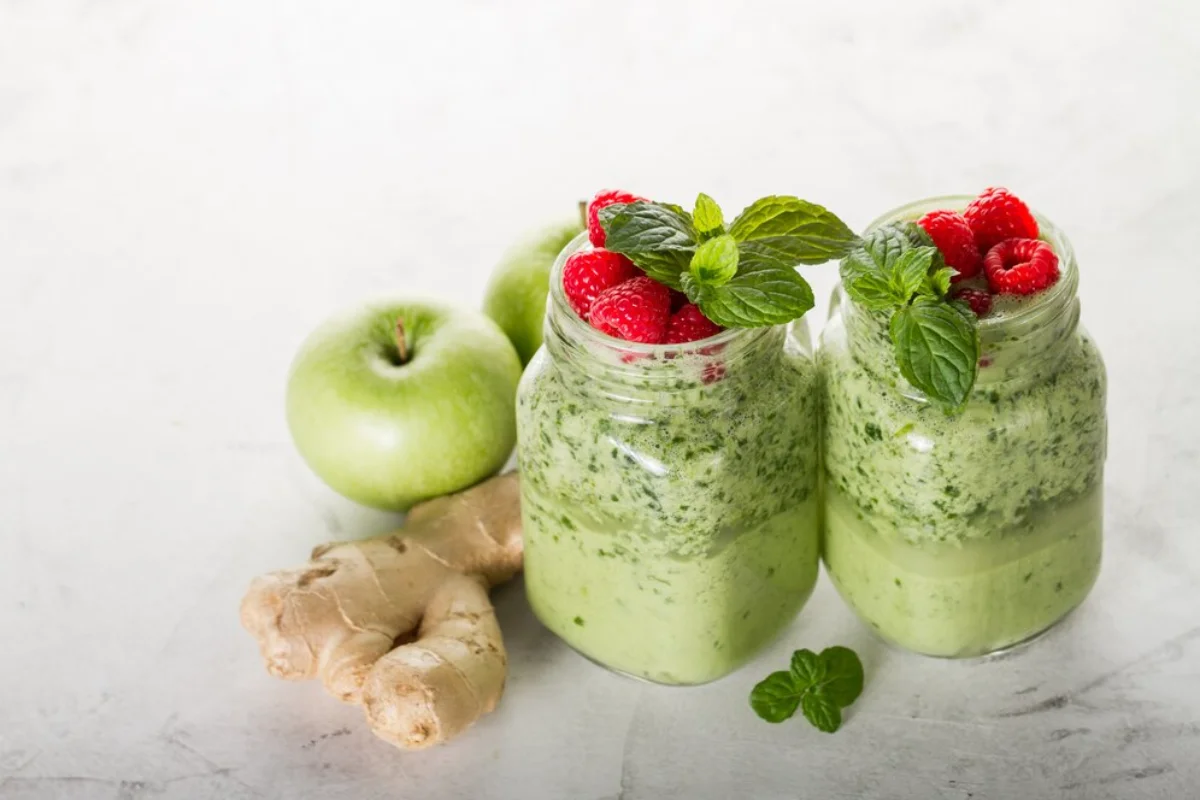 Two jars of green smoothie topped with fresh raspberries and mint leaves, accompanied by green apples and ginger