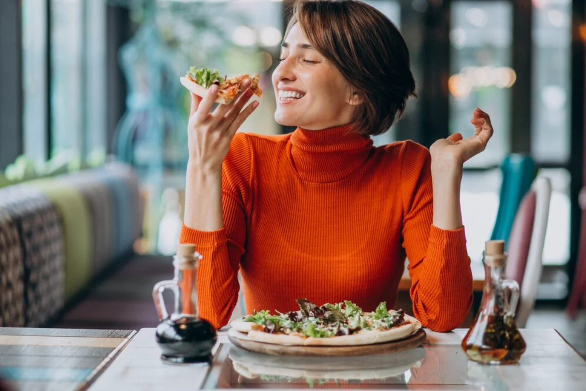 A woman in a bright orange turtleneck smiles with pleasure as she is about to eat a slice of pizza