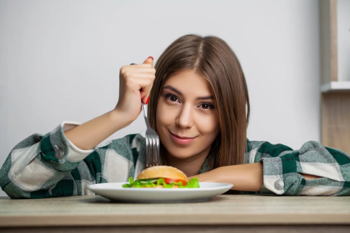 Young woman in a green plaid shirt smiling at the camera while sitting at a table, holding a fork, and about to eat a burger on a plate