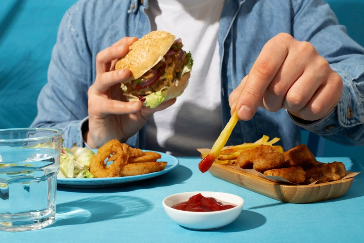 A person dips a chip into ketchup while holding a burger, with additional snacks like onion rings and fish fingers on a table