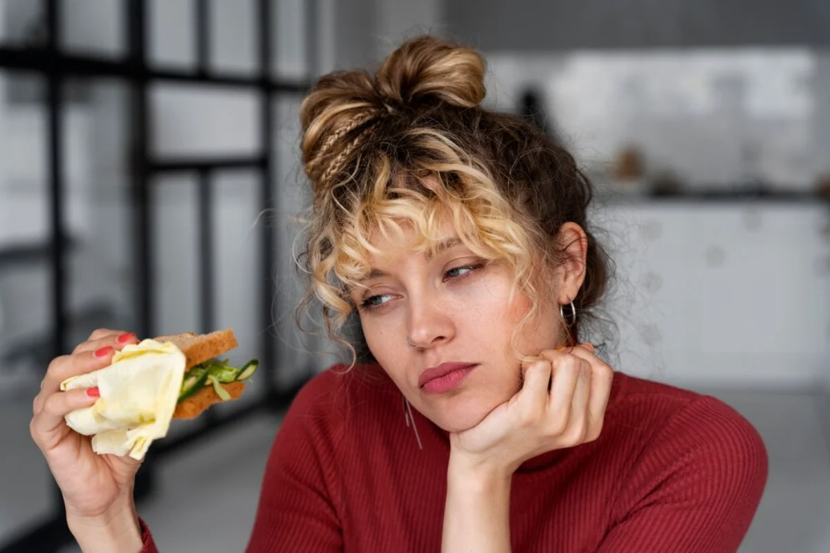 A pensive young woman with blonde curly hair tied up, holding a sandwich, sitting at a table in a modern kitchen.