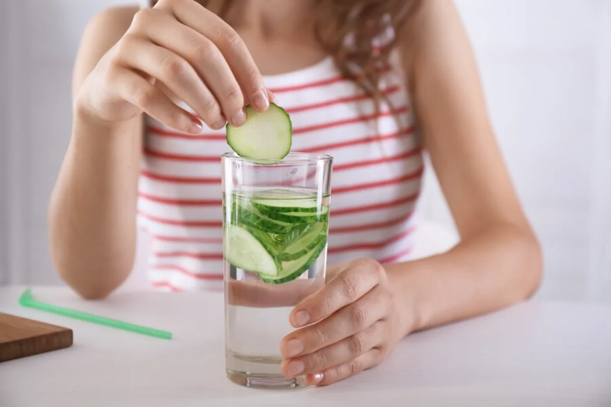 A woman in a striped top adding a slice of cucumber to a glass of water filled with cucumber slices.