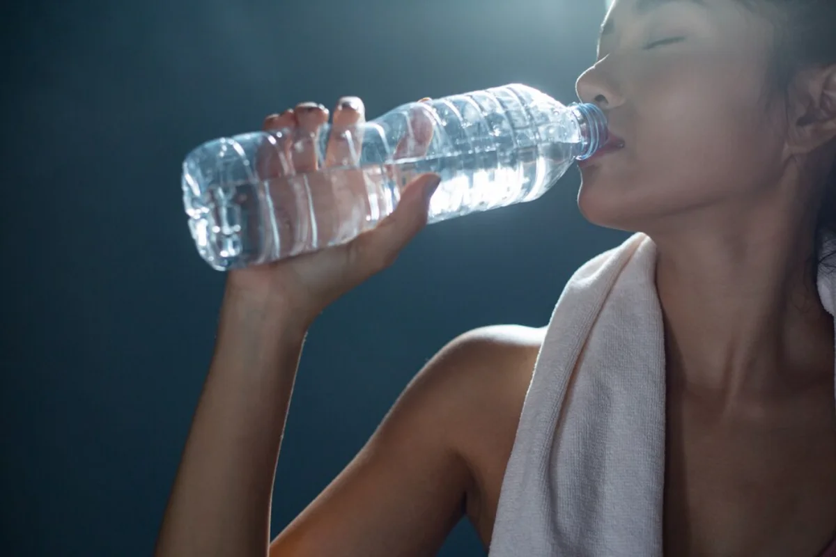 A woman with a towel around her neck drinks water from a clear plastic bottle, refreshing herself