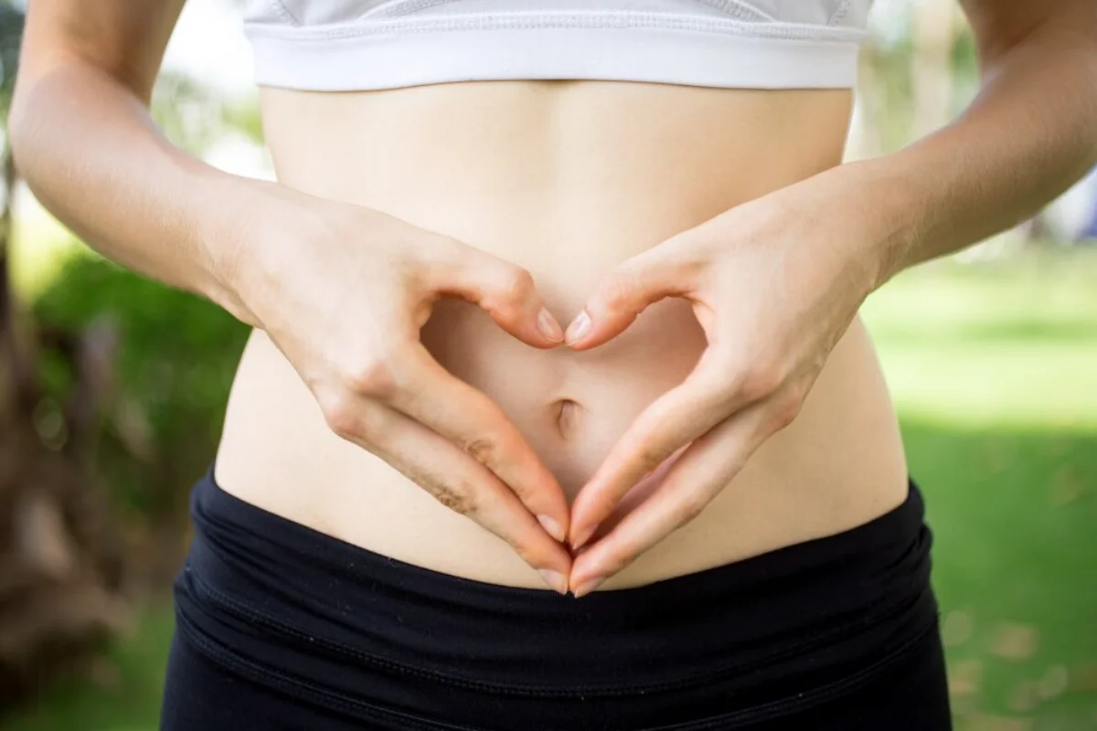 A close-up of a woman's midsection, where she is forming a heart shape with her hands around her belly