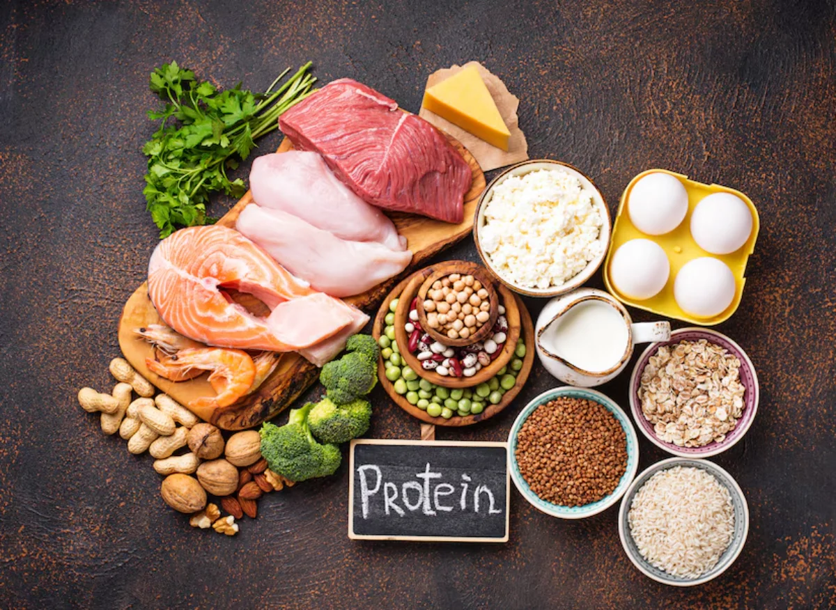 Assorted high-protein foods on a dark surface, including meats like beef and salmon, dairy products, eggs, nuts, and legumes