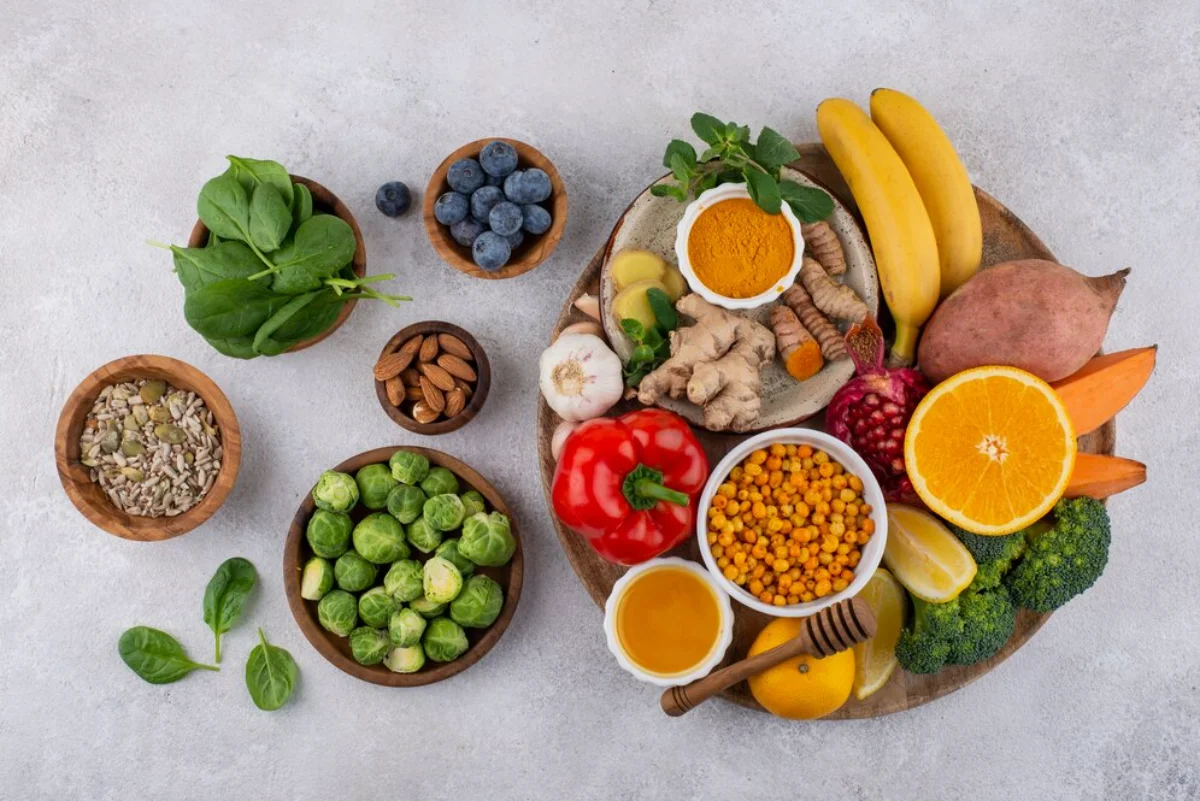 A variety of fresh vegetables, fruits, nuts, and seeds arranged on a pale surface