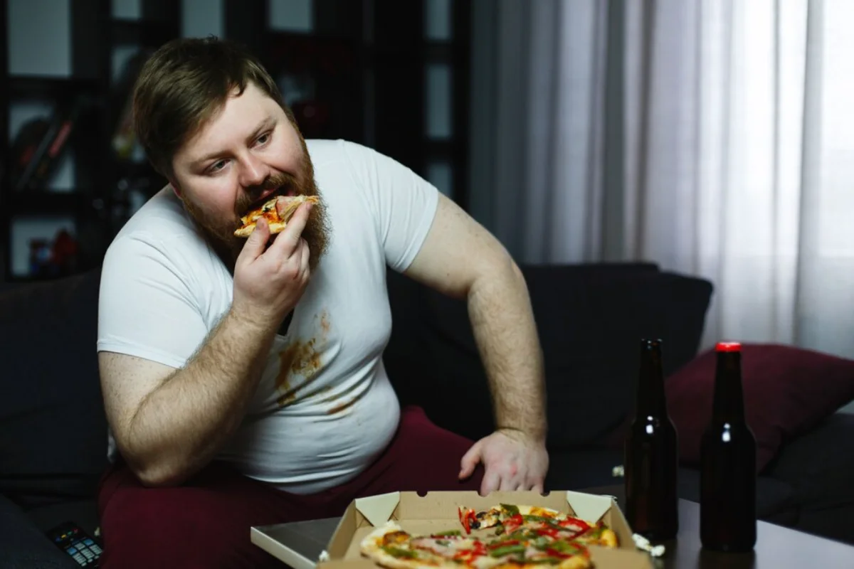 A man sitting on a sofa enjoying a slice of pizza, with a pizza box and two beer bottles