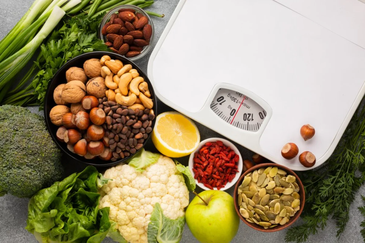 A variety of fresh vegetables and an assortment of nuts arranged next to a weighing scale on a kitchen countertop