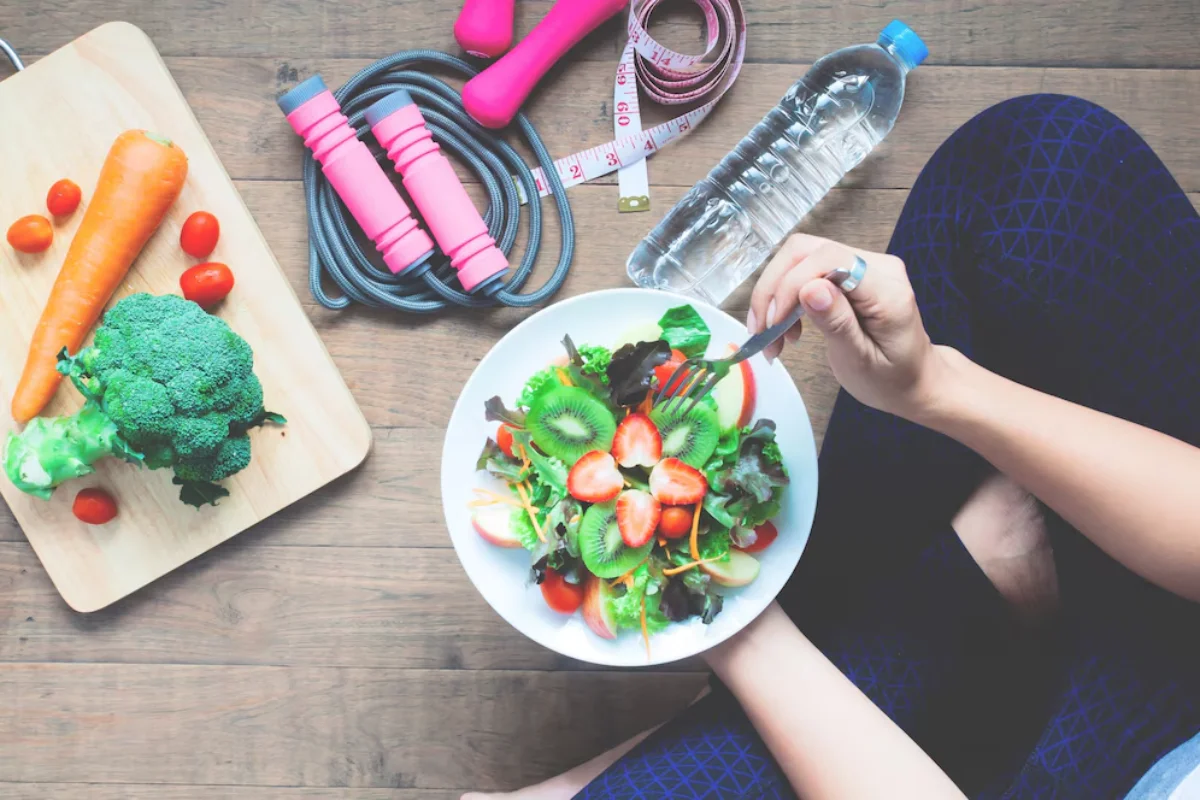 Top view of a person eating a colourful salad with kiwi, strawberries, and greens, next to workout gear including dumbbells