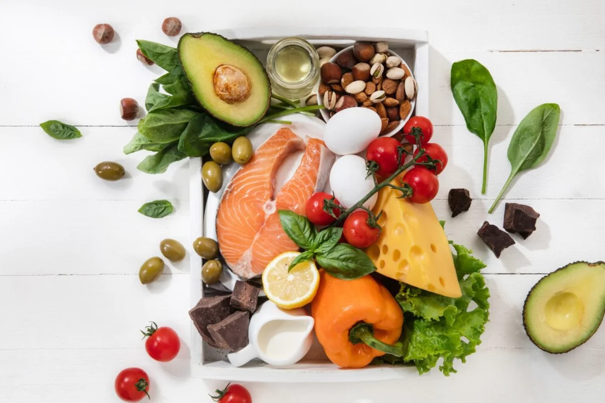 healthy foods including salmon, avocado, nuts, eggs, cheese, olives, dark chocolate, and fresh vegetables such as spinach, tomatoes