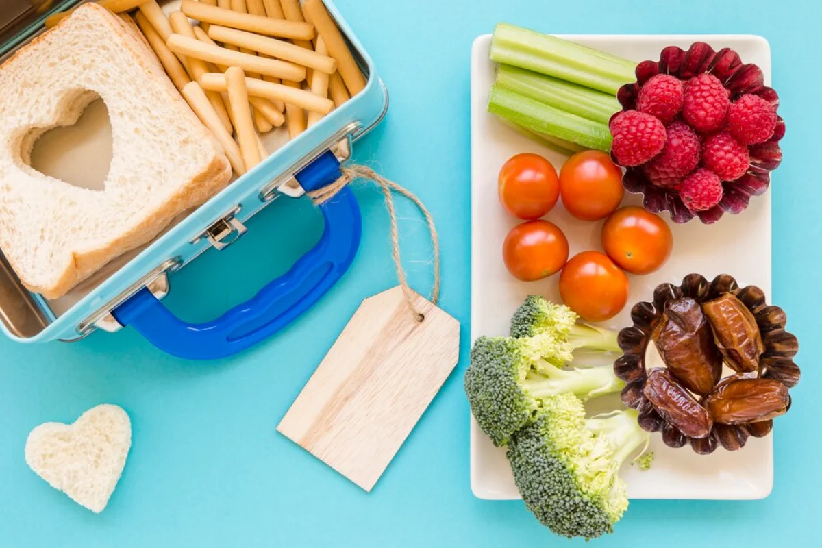 A nutritious meal setup on a turquoise background featuring a metal lunch box with a heart-shaped cut-out in a slice of bread and breadsticks