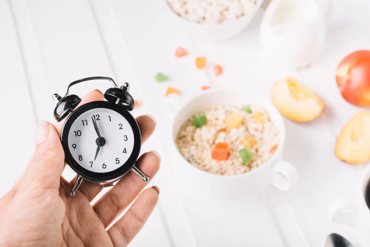 A person holding a small black alarm clock over a breakfast table with blurred bowls of oatmeal, fruits, and milk