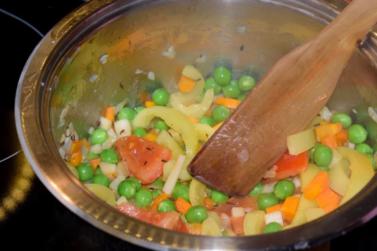 A mix of diced vegetables including peas, carrots, peppers, and onions being sautéed in a stainless steel pan with a wooden spoon.