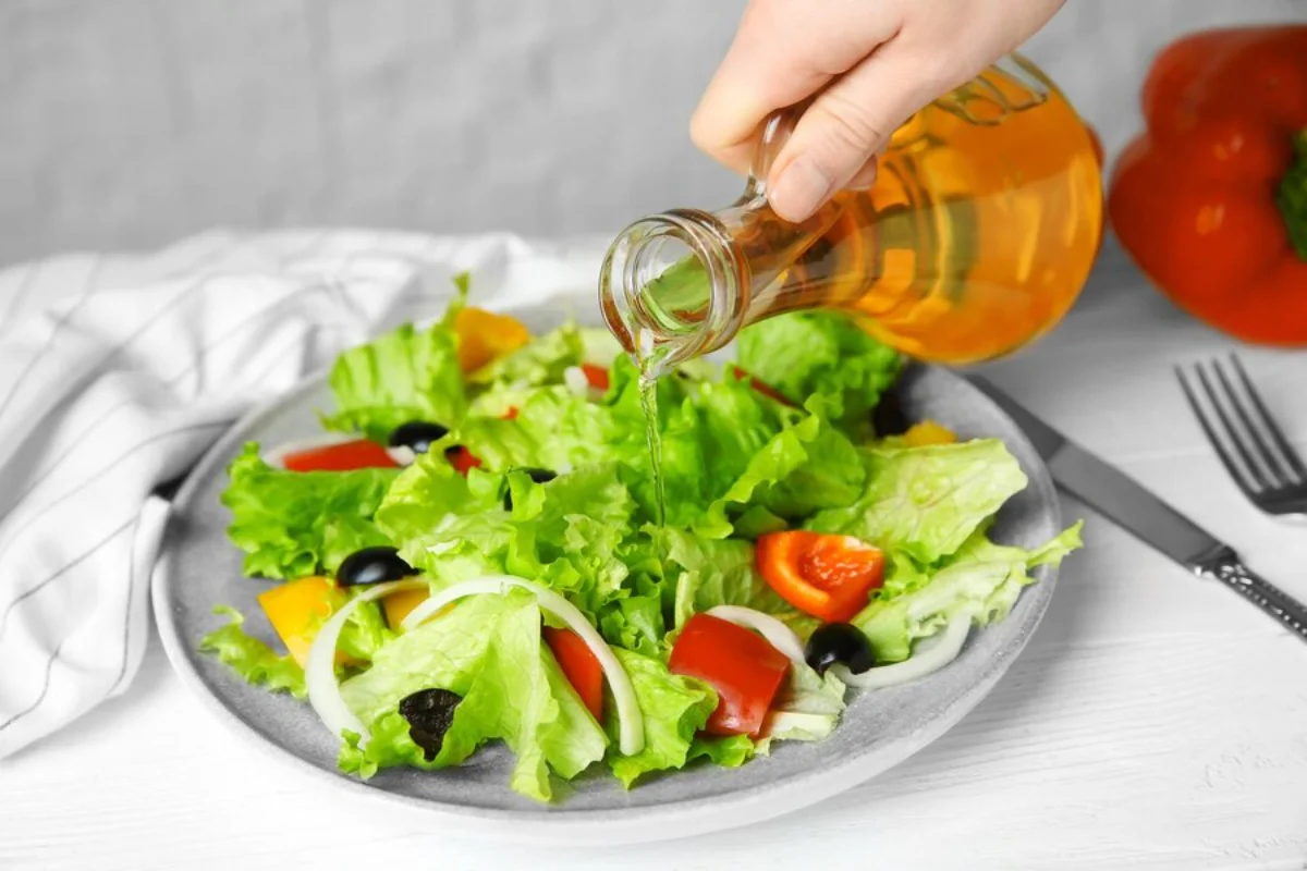 Drizzling olive oil onto a fresh salad with lettuce, tomatoes, peppers, onions, and olives on a grey plate,