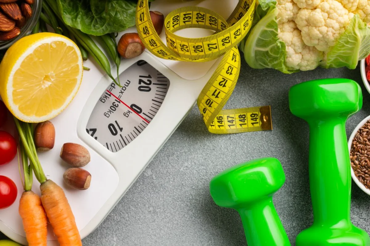 A top-down view of a weight loss and healthy living concept, featuring a variety of fresh vegetables, green dumbbells, and a bathroom scale