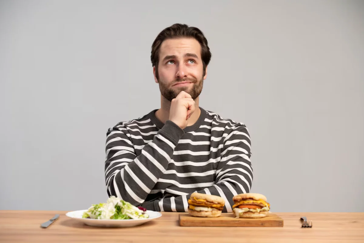 A man in a striped shirt sitting at a table, thoughtfully looking up while choosing between a salad and hamburgers.