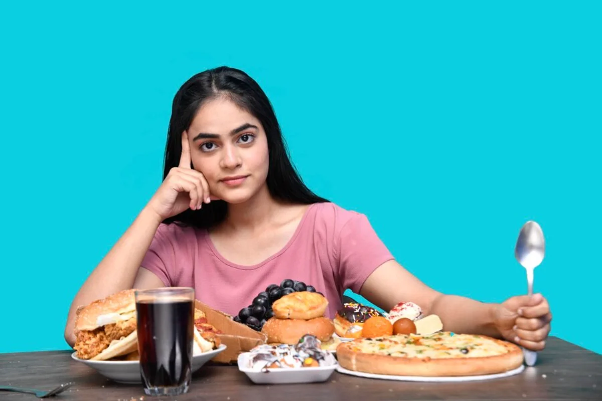 Young woman sitting at a table with various unhealthy foods, looking contemplative