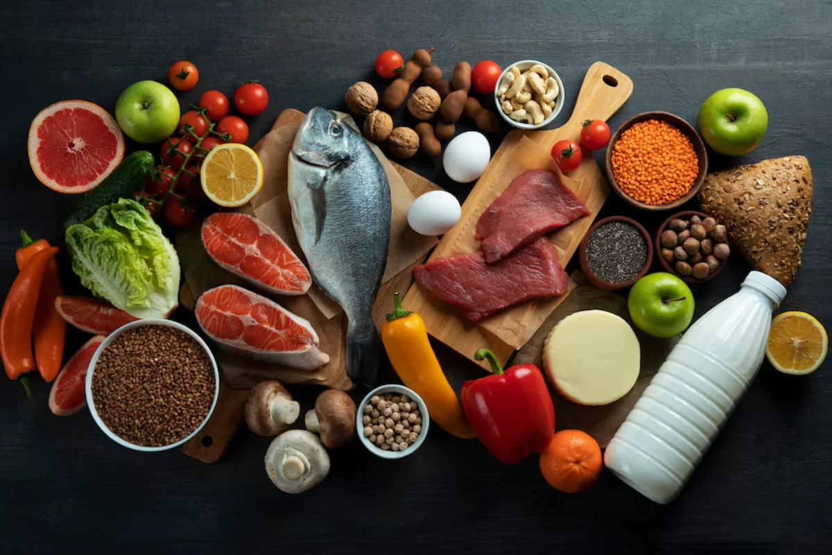 A variety of healthy foods displayed on a dark wooden surface including fruits, vegetables, nuts, whole grains, seafood, meats, and dairy.