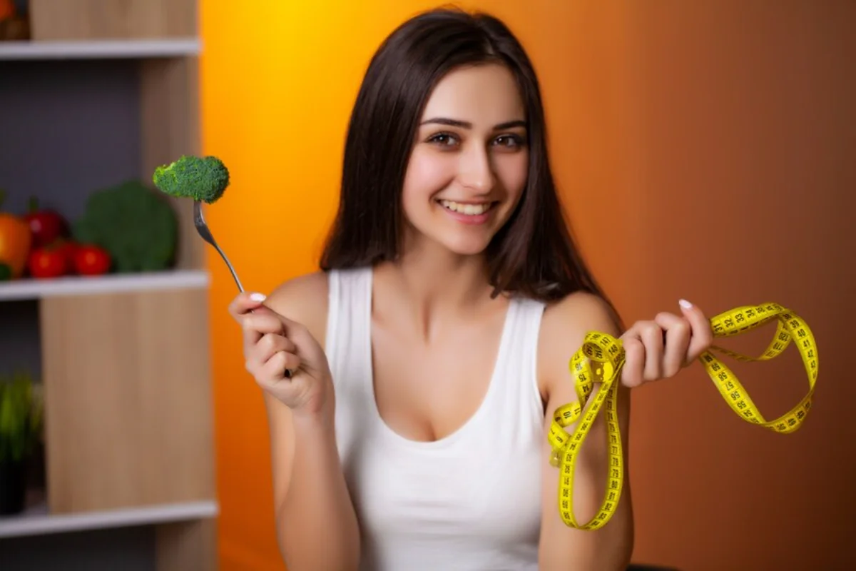 A smiling young woman in a white tank top holding a fork with broccoli and a measuring tape, promoting healthy eating and fitness