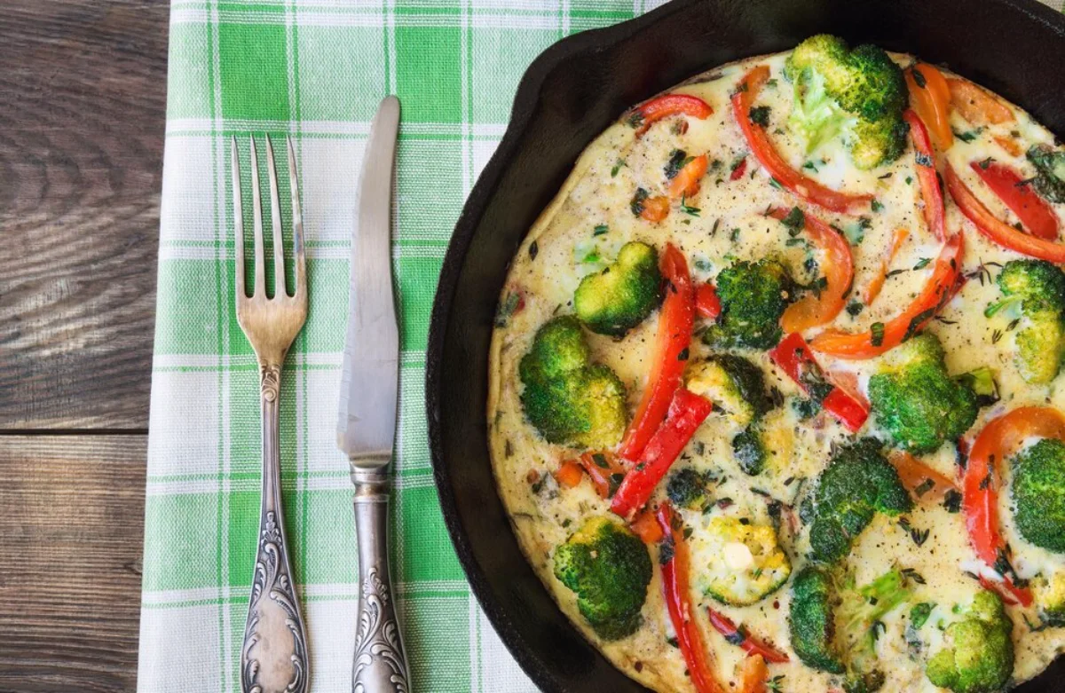 A vibrant vegetable frittata in a cast iron skillet on a wooden table, featuring broccoli, red peppers, and herbs