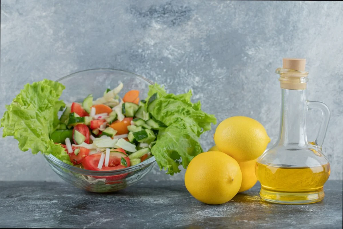 A fresh vegetable salad in a glass bowl, alongside two lemons and a bottle of olive oil 