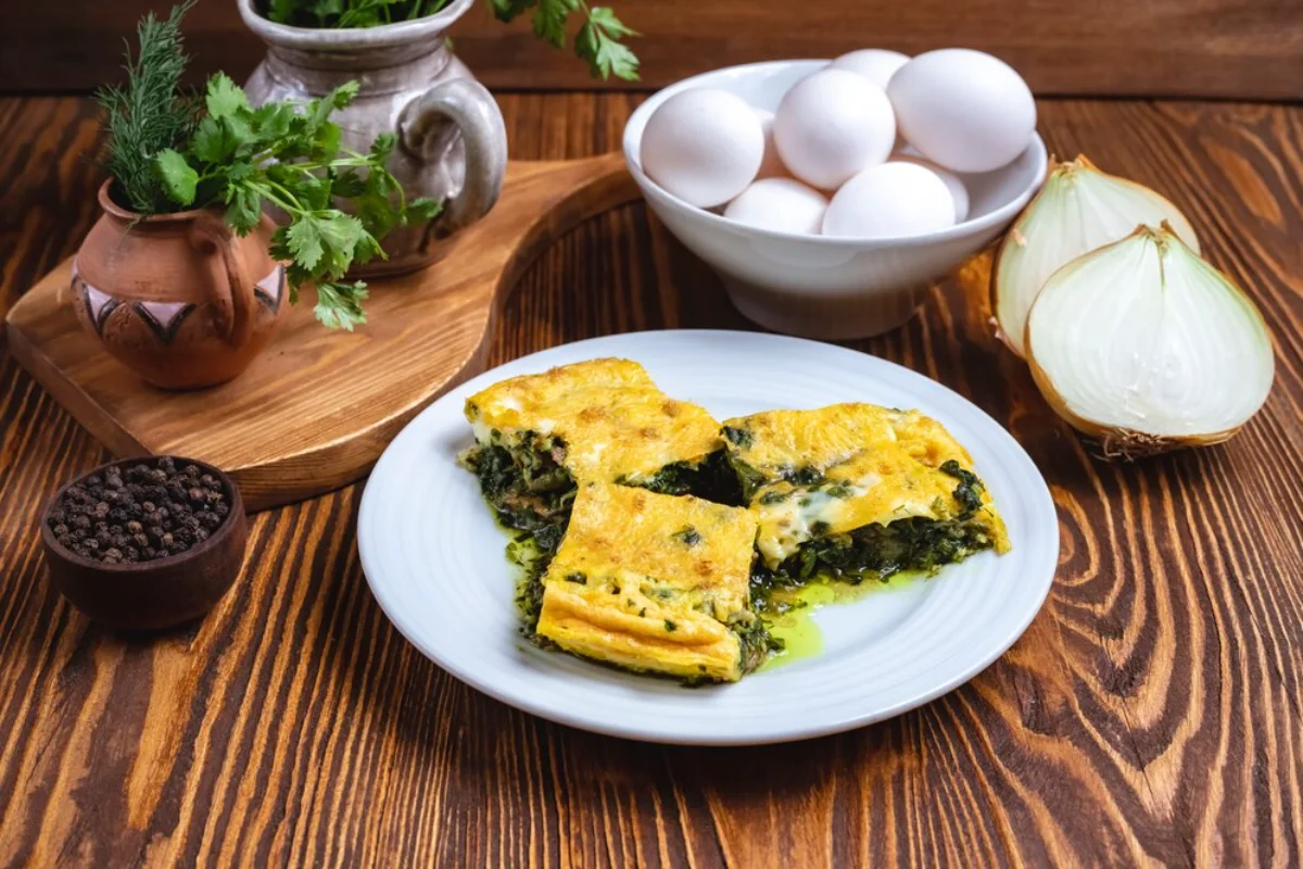 Slices of spinach omelette on a white plate with fresh herbs in a terracotta pot, peppercorns in a small bowl