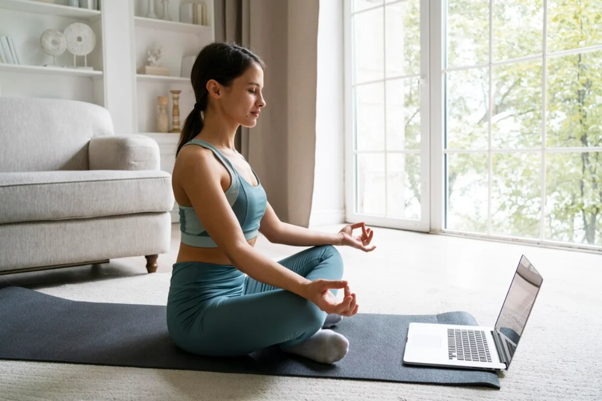 A woman in a teal sports bra and leggings meditates in a seated position on a yoga mat 