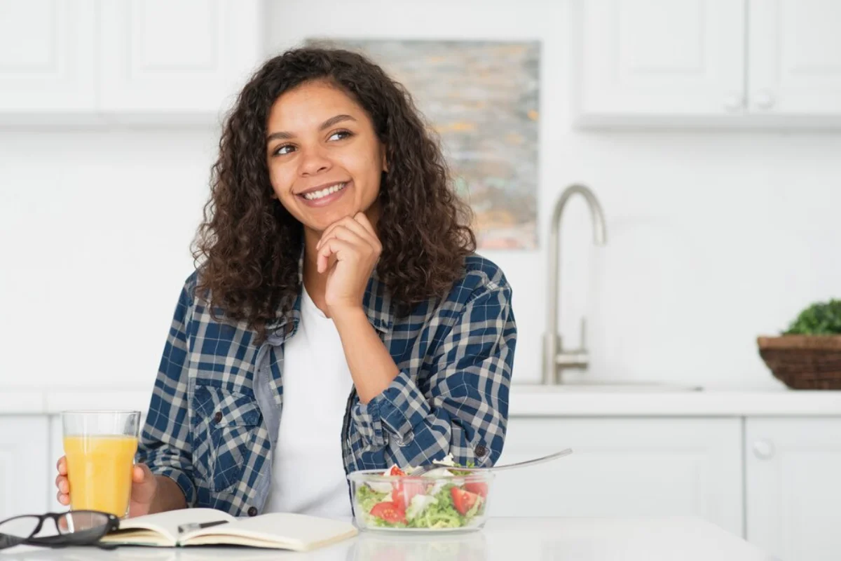 A young woman smiling, seated at a kitchen table with a bowl of salad, a glass of orange juice, and an open book in front of her