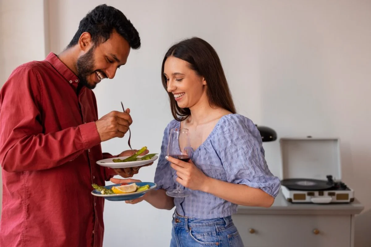 A man and a woman enjoying a meal together in a home setting, with the man sampling food from a plate while the woman holds a glass of wine