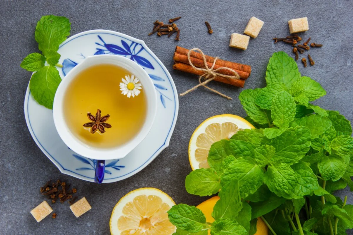 A cup of herbal tea with a star anise and a small daisy floating in it, surrounded by fresh mint leaves