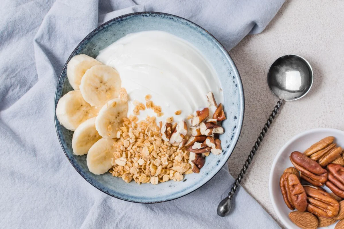 A bowl of yogurt garnished with sliced bananas, granola, and chopped nuts, accompanied by a silver spoon and a small bowl of pecans and almonds