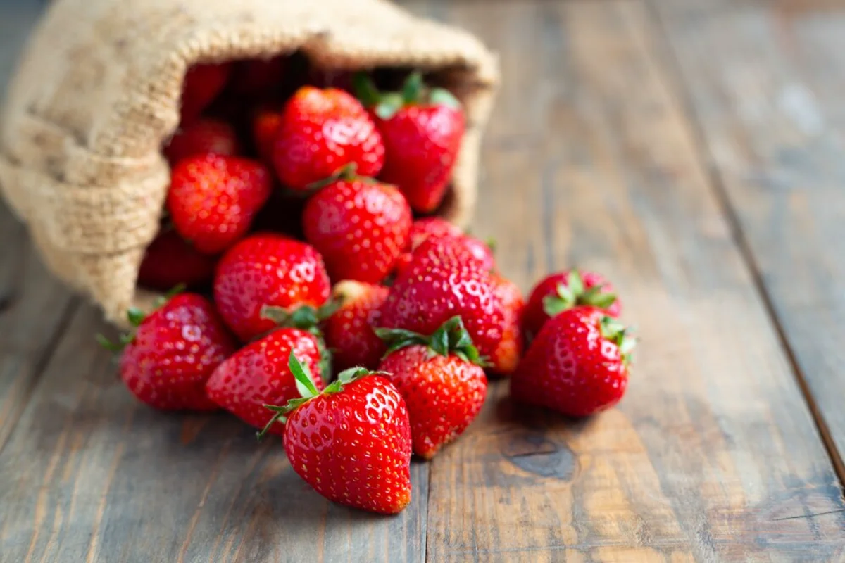 A collection of fresh, vibrant strawberries spilling out from a burlap sack onto a rustic wooden surface.