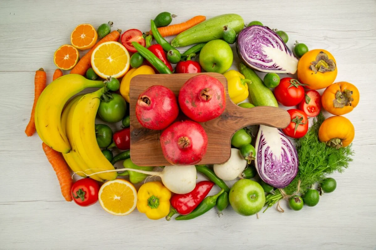 A variety of fresh fruits and vegetables spread out on a light wood surface, including bananas, oranges, carrots, tomatoes
