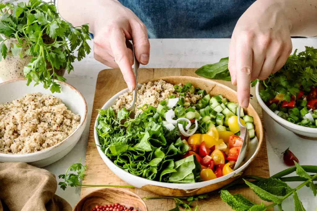 A person's hands assembling a vibrant and fresh vegetable and grain salad with various ingredients like lettuce, tomatoes, cucumbers