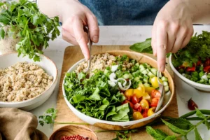 A person's hands assembling a vibrant and fresh vegetable and grain salad with various ingredients like lettuce, tomatoes, cucumbers