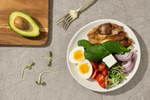 healthy meal on a grey fabric surface, featuring a plate with bacon, boiled egg halves, spinach leaves, sliced red onions, cherry tomatoes