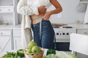 A person measuring their waist with a measuring tape in a bright kitchen, with fresh green fruits and vegetables