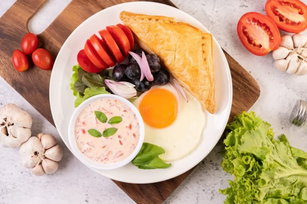 A plate of breakfast food featuring a sunny-side-up egg, a golden-brown pastry, a bowl of pink sauce with herbs, tomato slices