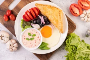 A plate of breakfast food featuring a sunny-side-up egg, a golden-brown pastry, a bowl of pink sauce with herbs, tomato slices