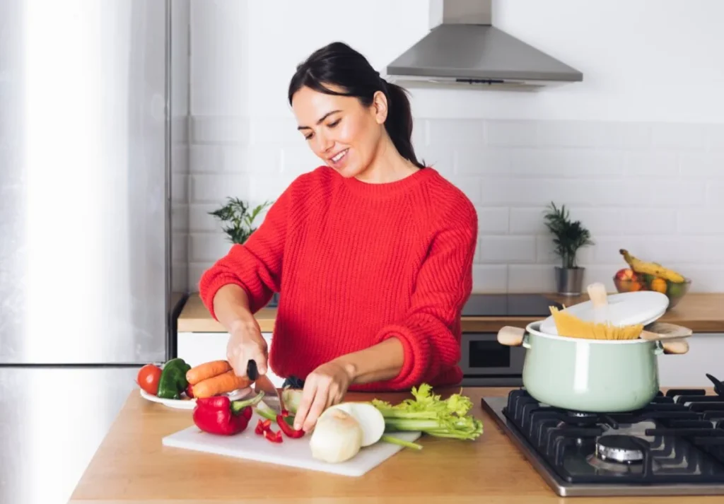 A woman in a red sweater smiling while chopping vegetables in a modern kitchen.