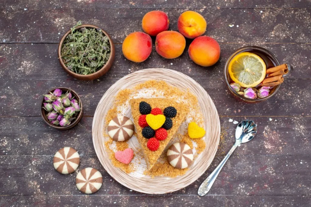 a cake slice decorated with colourful fruit berries in a geometric pattern, surrounded by fresh apricots, a bowl of tea leaves