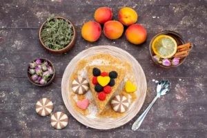 a cake slice decorated with colourful fruit berries in a geometric pattern, surrounded by fresh apricots, a bowl of tea leaves