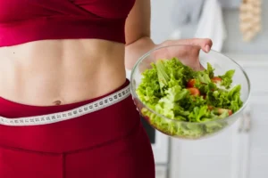 A close-up of a woman in a red sports top, holding a tape measure around her waist and a bowl of salad in her hand