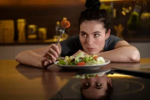 A woman with a contemplative expression sits at a table, eating a salad, with her reflection