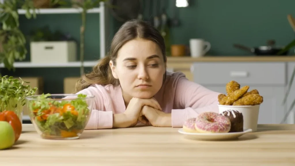 A woman in a pink top sits at a kitchen table, looking longingly at a plate of doughnuts and a cup containing fried snacks, with a bowl of salad nearby,